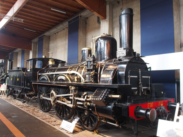 Alte Dampflokomotive in einem Museums Setting mit umliegenden Eisenbahnschienen, Informationsschildern und anderen Ausstellungen, vor einer Wand mit Deckenbeleuchtung.