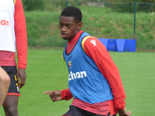 Ein junger Mann in einem blauen Hemd und schwarzen Shorts spielt Fußball auf einem grünen Feld, das von einem Zaun, Pflanzen und Bäumen umgeben ist und scheint mitten in einem Training zu sein.