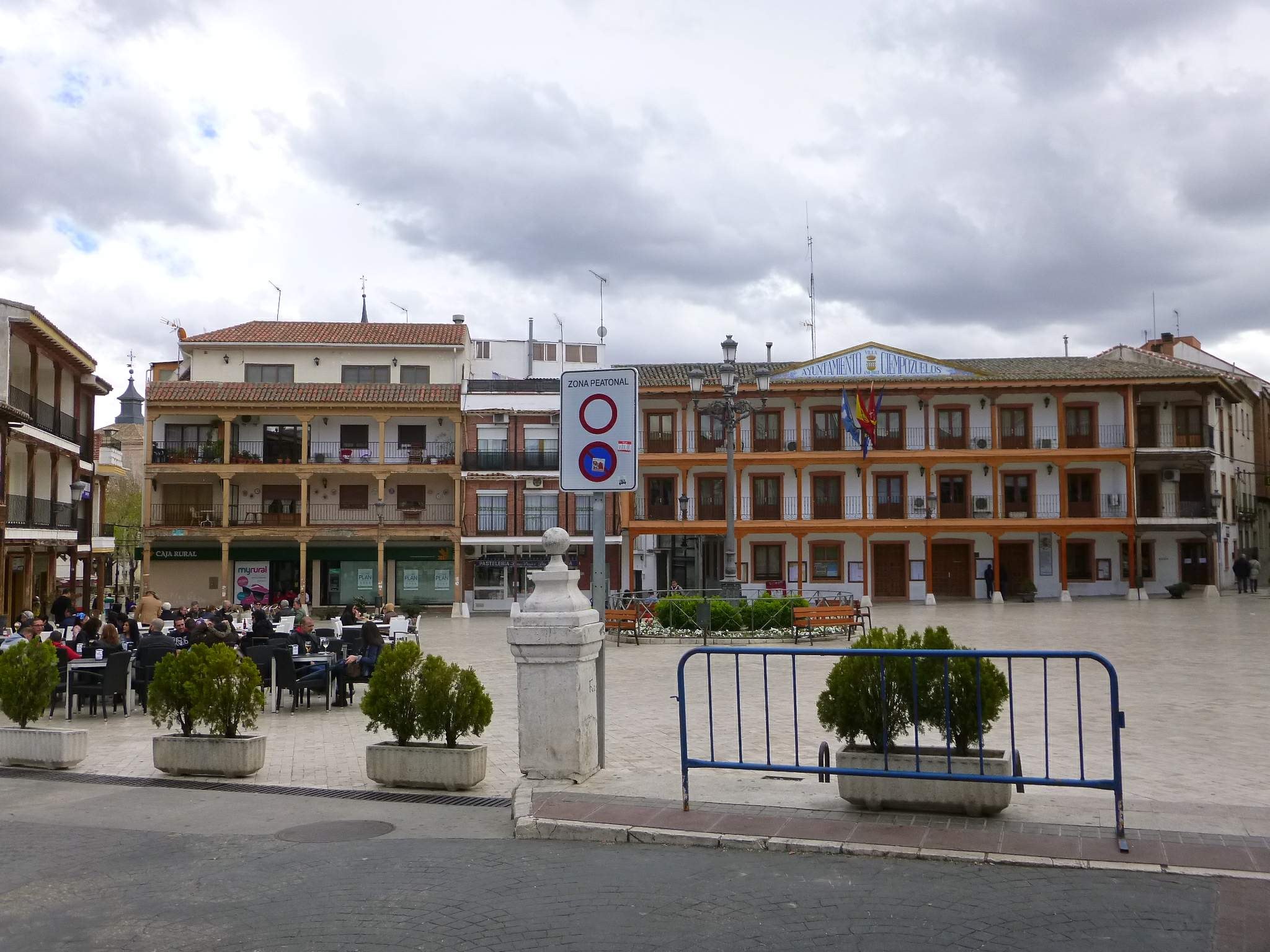Ein belebter Stadtplatz mit Menschen, die sitzen und stehen, Topfpflanzen, eine Metallabsperrung, ein Schild an einem Pfahl, Straßenlaternen mit Flaggen, umgebende Gebäude und ein bewölkter Himmel.