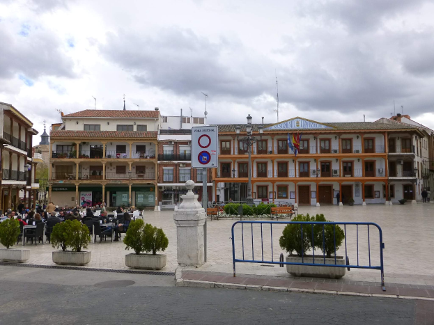 Ein belebter Stadtplatz mit Menschen, die sitzen und stehen, Topfpflanzen, eine Metallabsperrung, ein Schild an einem Pfahl, Straßenlaternen mit Flaggen, umgebende Gebäude und ein bewölkter Himmel.