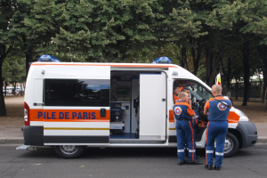 Ambulanz am Straßenrand geparkt mit drei Personen davor, Bäume und Gebäude im Hintergrund.