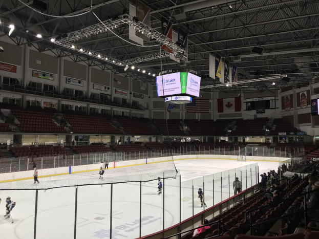 Eishockeyspiel in einer großen Arena mit einem zentralen Netz, Zuschauern auf beiden Seiten, Deckenleuchten und Trägern sowie Bannern an den Wänden, die 'St. Louis Blues Arena' beschriftet sind.