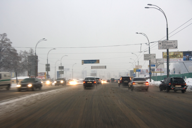 Eine belebte Stadtstraße an einem verschneiten Tag mit fahrenden Fahrzeugen, schneebedeckter Straße, Laternen, Texttafeln, Bäumen, Gebäuden und einem Himmel im Hintergrund.