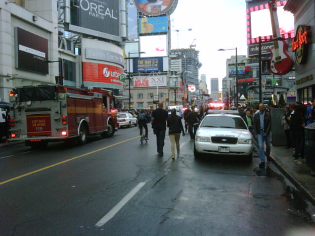 Eine belebte Straße mit parkenden Fahrzeugen auf beiden Seiten, Menschen, die auf der Straße und den Gehwegen gehen, und Geschäfte auf beiden Seiten.