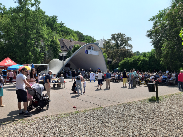 Eine Menschenmenge bei der Festivalöffnung, einige sitzen auf Bänken, andere stehen, mit einem Kinderwagen im Vordergrund, Zelte links, ein Müllimer rechts und Gebäude und Bäume im Hintergrund unter einem klaren blauen Himmel.