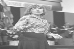 Eine Frau in einem Schwarz-Weiß-Foto steht auf einer Bühne, trägt ein weißes Hemd und einen schwarzen Rock und hat ihr Haar zu einem Dutt gebunden, umgeben von einer Gruppe von Menschen und einer Wand im Hintergrund.