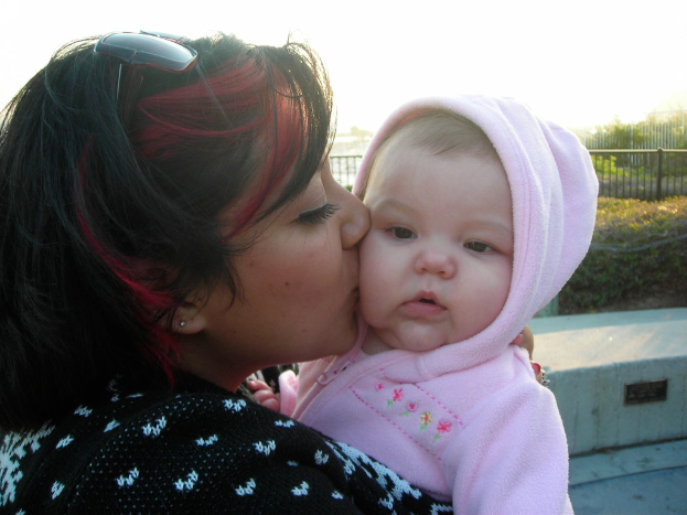 Eine Frau umarmt ein Baby in der Mitte des Bildes, mit Büschen und Zaun im Hintergrund und Boden sichtbar in der rechten unteren Ecke.