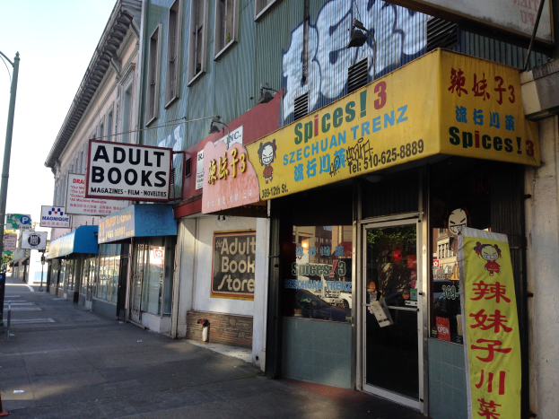 Eine belebte Stadtstraße mit verschiedenen Gebäuden, Fahrzeugen und einem Schaufenster, das ein Schild trägt, auf dem "Erwachsene Bücher" steht, unter einem klaren blauen Himmel.