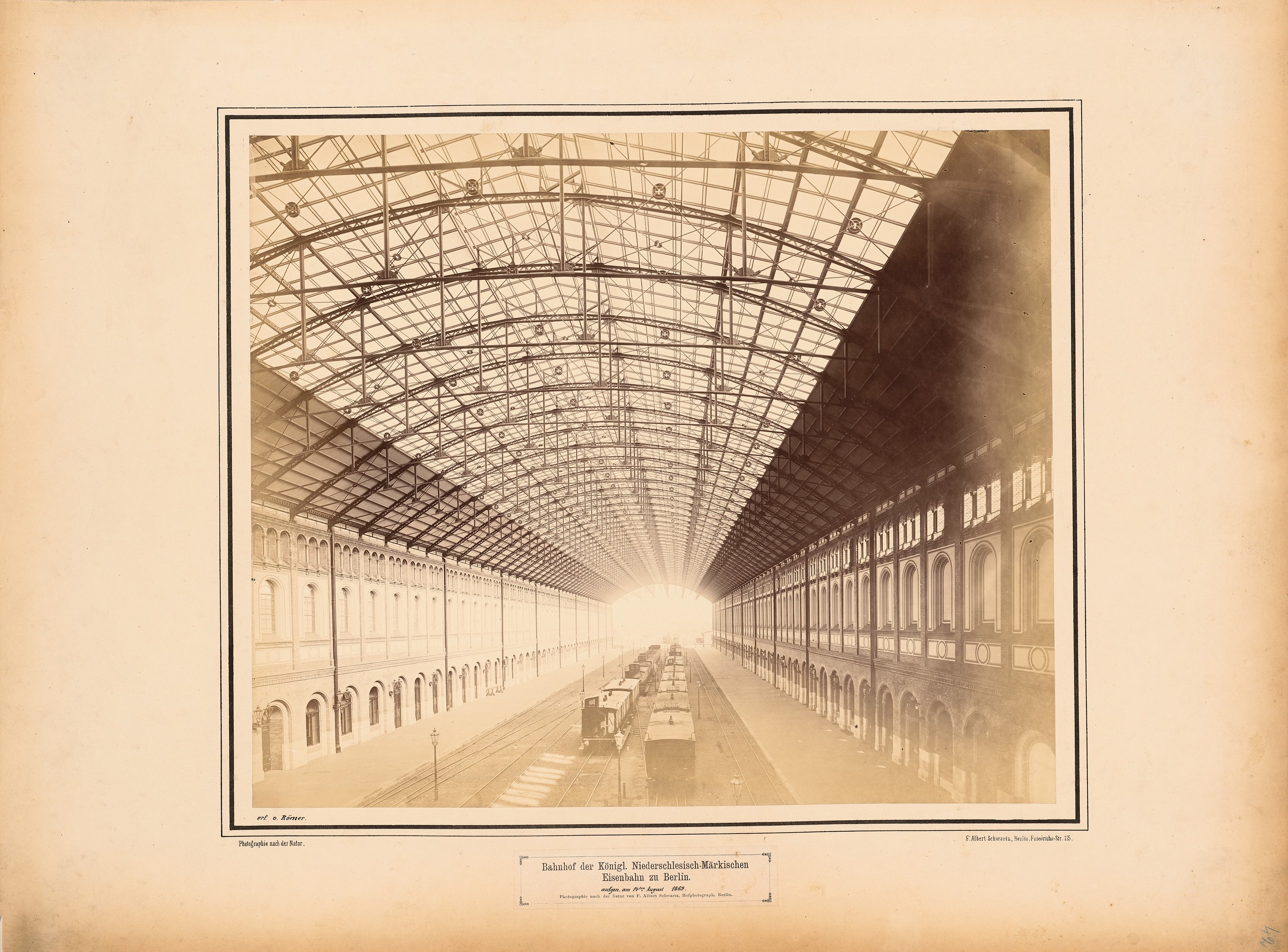 Schwarz-weiß-Foto eines Bahnhofs mit einem Zug auf den Schienen, umgeben von Gebäuden mit Fenstern und einem Dach, das von Metallpfosten gestützt wird; Text ist unten sichtbar.