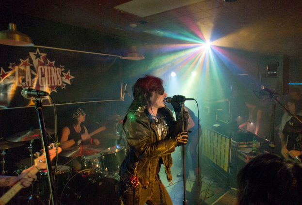 Eine Frau in einer schwarzen Jacke singt auf der Bühne in ein Mikrofon, mit einem Mann in Sonnenbrille und einem Stirnband, der hinter ihr ein Instrument spielt, und einem Plakat mit der Aufschrift "LA Guns" im Hintergrund, unter Disco- und Hängelampen.