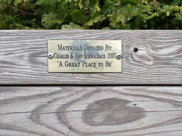 Holzbank mit einer Tafel, auf der "Materialien gespendet von Charles & Ann Schwabach 2017: Ein toller Ort" steht, vor üppigen grünen Pflanzen.