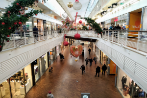 Ein Einkaufszentrum im Weihnachtslook mit Menschen, die herumlaufen, Schaufensterpuppen in Festkleidung, Geschäften, Geländern und Lichtern, die eine gemütliche Atmosphäre schaffen.