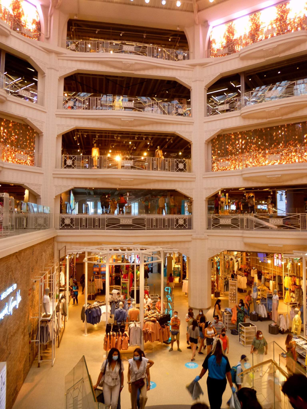 Ein belebter Einkaufszentrumsvorplatz mit Menschen, die Taschen tragen und Schaufensterpuppen mit Kleidern, umgeben von Geländern, Lampen und Texttafeln an den Wänden.