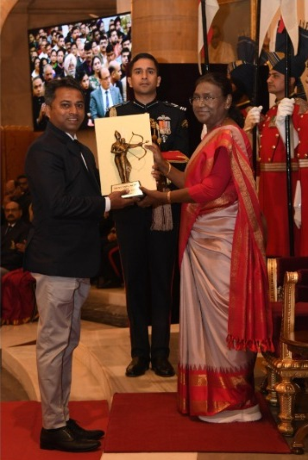 Ein Mann in einem schwarzen Blazer, weißem Hemd und grauen Hosen hält eine Trophäe neben einer Frau in einem ashesfarbenen Sari auf einem roten Teppich bei einer Preisverleihung, mit Menschen, Stühlen, Fahnen und einem Bildschirm im Hintergrund.