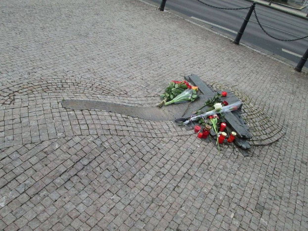 Ein Straßenrand-Denkmal mit Blumen, Kerzen und verstreuten Buketts vor einem kleinen Denkmal, umgeben von Kettenzaun-Pfählen und vorbeifahrenden Fahrzeugen, mit Bäumen und Gebäuden im Hintergrund.