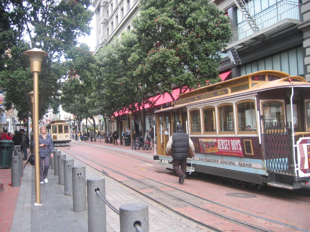 Schienen verlaufen entlang einer Straße mit einem Zug, der Text anzeigt, einer Person in der Nähe, kleinen Pfosten mit Seilen auf der linken Seite, Straßenlaternen, Bäumen, vielen Menschen im Hintergrund, einem Müllcontainer auf der linken Seite und Gebäuden mit Leitern auf der rechten Seite.