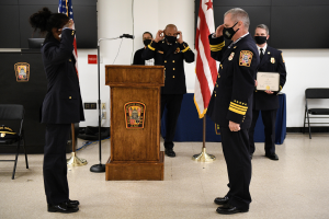 Eine Gruppe uniformierter und maskierter Personen steht vor einem Podium mit einem Mikrofon, umgeben von Flaggen, Stühlen und Tischen, während eine Vereidigung für einen neuen Feuerchef stattfindet, wobei eine Person ein Zertifikat hält.