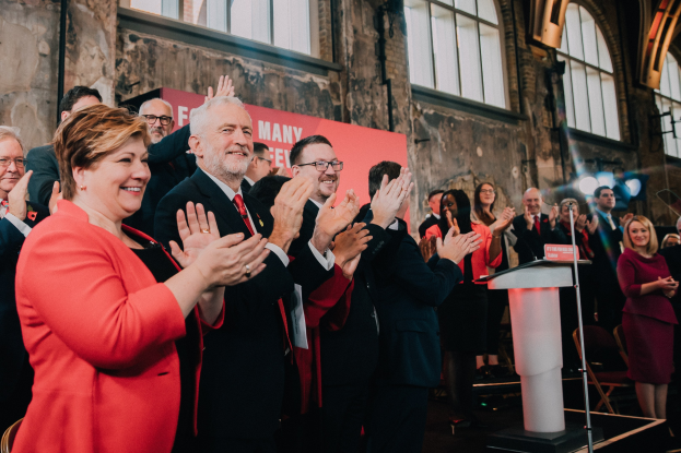 Eine Gruppe von Menschen, die vor einem Publikum klatschen, mit einem Podium, einem Mikrofon und einer Tafel mit Text auf der rechten Seite, sowie Stühlen, einer Fahne, einer Wand, Fenstern und Lichtern im Hintergrund.