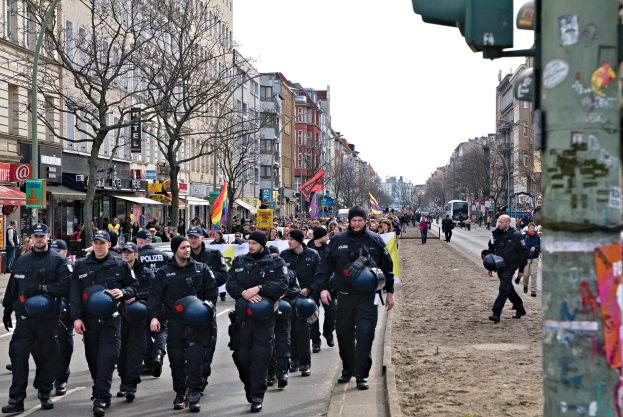 Polizeibeamte in schwarzen Uniformen und Helmen gehen eine von hohen Gebäuden gesäumte Stadtstraße entlang, einige tragen Fahnen, mit einem mit Aufklebern bedeckten Pfahl auf der rechten Seite und Bäumen, Laternenmasten und einem klaren blauen Himmel im Hintergrund.