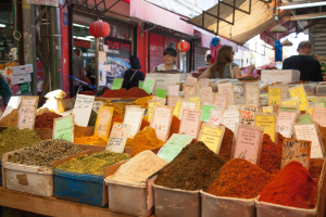Ein belebter Markt mit verschiedenen Gewürzkisten, jede mit einem Preisetikett, Menschen im Hintergrund und ein Stand mit Ausstellungsstücken auf der linken Seite, alles von Deckenleuchten beleuchtet.