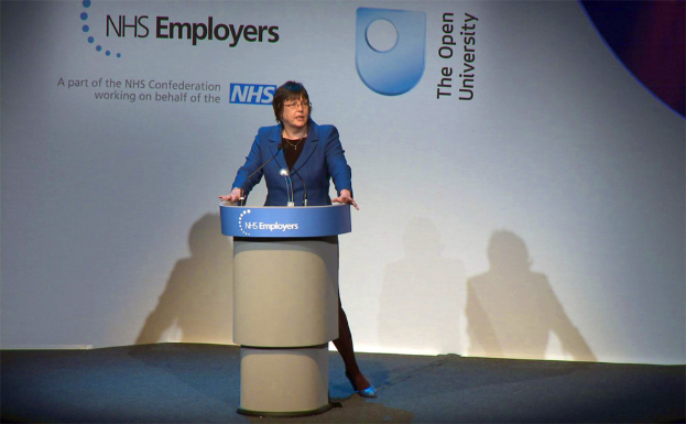 Eine Frau steht an einem Podium mit Mikrofonen, trägt Kleidung, geschnittene Schuhe und eine Brille, mit einem Plakat und ihrem Schatten dahinter.