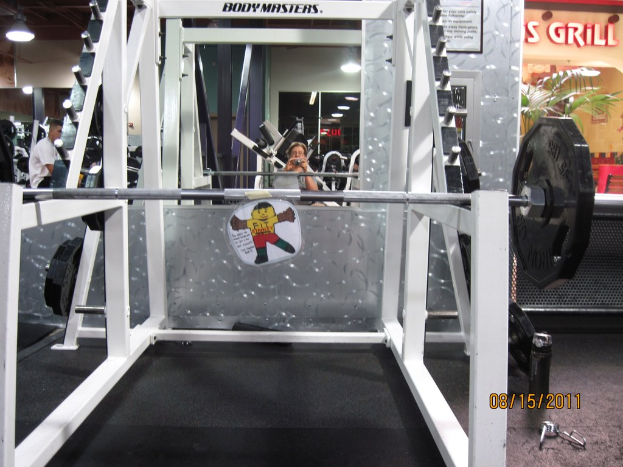 Ein Fitnessstudio mit einer Hantelbank im Vordergrund, einem Mann links und einer Frau in der Mitte.