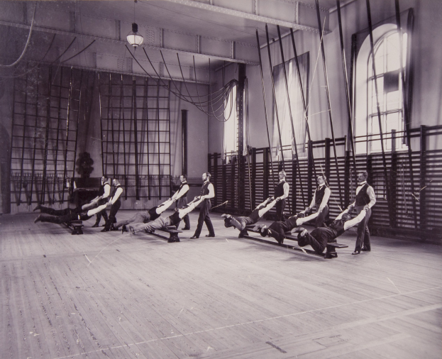 Schwarzes und weißes Bild einer Gruppe von Menschen, die Pilates-Übungen in einem Fitnessstudio mit einer Wand, Fenstern auf der rechten Seite und einer Decke mit Leuchten im Hintergrund durchführen.