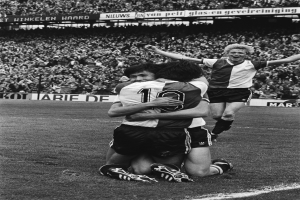 Drei Fußballspieler umarmen sich auf dem Boden, feiern ein Tor, mit jubelnder Menge und schwenkenden Fahnen im Hintergrund, dargestellt in Schwarz-Weiß.