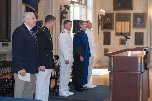 Gruppe von Männern in einem formellen Raum mit einem Podium, Mikrofon, Stühlen, Tisch, Flagge, Foto Rahmen, Kerzen und einer Tür.