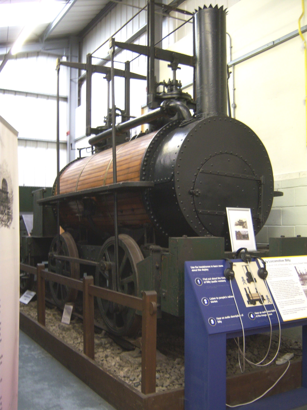 Alte Dampflokomotive in einem Museum mit Informationsschildern, einem Zaun, Steinen, einer Fahne, einer Wand mit Rohren und Deckenbeleuchtung ausgestellt.
