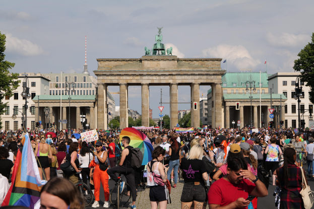 Eine große Menschenmenge marschiert vor dem Reichstagsgebäude in Berlin, Deutschland, viele halten Fahnen und Schilder, einige fahren Fahrräder, mit Laternenmasten, Bäumen und bewölktem Himmel im Hintergrund und Gebäuden mit Fenstern, Bögen und einer Statue auf dem Bogen.