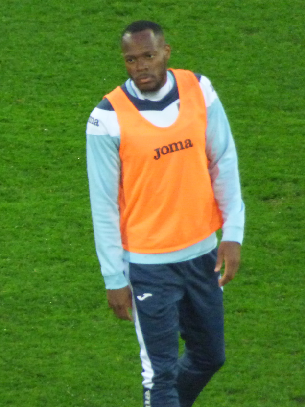 Ein Mann in einem orangen T-Shirt und blauen Hosen, der wie ein Fußballspieler aussieht, läuft entschlossen über ein saftiges grünes Feld.