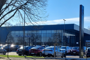 Ein Mercedes-Autohaus mit parkenden Autos davor, umgeben von einem Metallzaun, Gras, einem Schild, Straßenlaternen, einer Gruppe von Bäumen und einem bewölkten Himmel.
