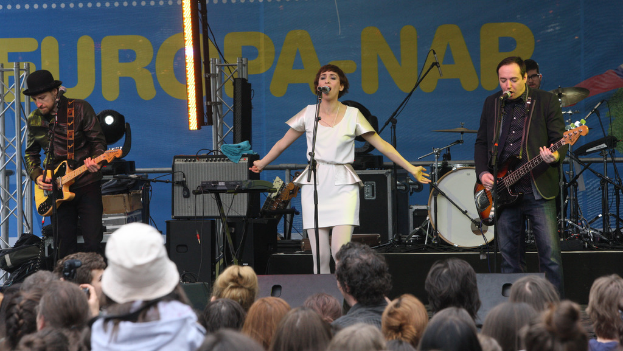 Ein Konzertauftritt mit vier Personen auf der Bühne: eine Frau in einem weißen Kleid in der Mitte, zwei Männer mit Gitarren links und rechts daneben, eine Menge unten und Lautsprecher im Hintergrund.