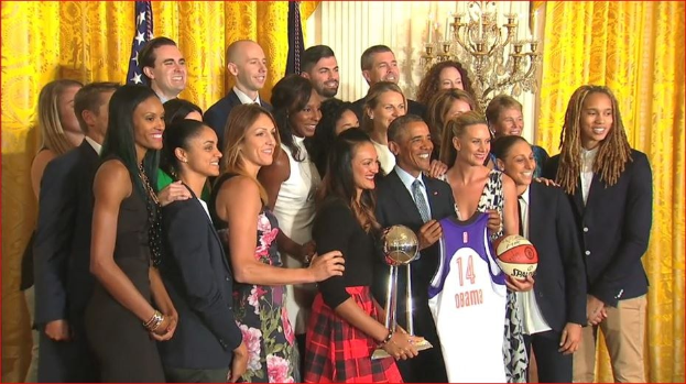Präsident Obama und First Lady Michelle Obama posieren mit dem Damen-Basketball-Team im Oval Office des Weißen Hauses, halten einen Basketball, eine Trophäe und lächeln in der Nähe einer Flagge, Vorhänge und einer Kerzenleuchter.
