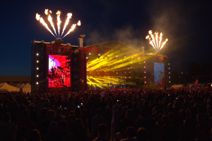Eine große Menschenmenge auf einem Musikfestival mit einer Bühne, auf der Künstler unter hellen Scheinwerfern auftreten und Banner schmücken den Hintergrund, Zelte links und ein Feuerwerk, das den dunklen Himmel erhellt.
