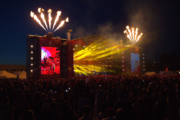 Eine große Menschenmenge auf einem Musikfestival mit einer Bühne, auf der Künstler unter hellen Scheinwerfern auftreten und Banner schmücken den Hintergrund, Zelte links und ein Feuerwerk, das den dunklen Himmel erhellt.