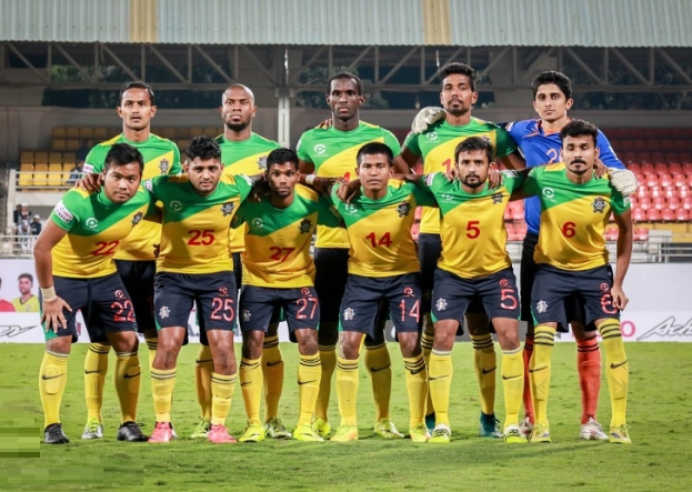 Eine Gruppe von Männern auf einem Fussballfeld, umgeben von Werbetafeln, Stühlen, Geländern, Pfosten, Bäumen und einer Hütte im Hintergrund, scheinen einen Sieg zu feiern.