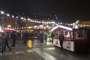 Ein belebter Weihnachtsmarkt bei Nacht in einer Stadt, mit Menschen, die spazieren gehen, Ständen mit Lichtern und Dekorationen, Gebäuden mit Fenstern, Bäumen und festlichen Lichtern am Himmel.
