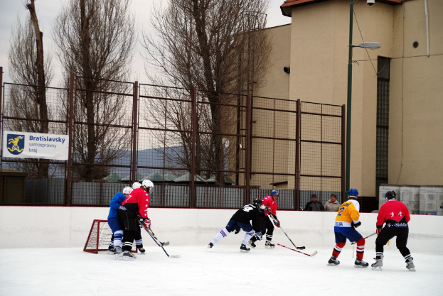 Personen beim Eishockeyspielen auf einem Eisring mit Gebäuden, Bäumen, einer Straßenlaterne, einem Namensschild und Zäunen im Hintergrund unter einem Himmel.