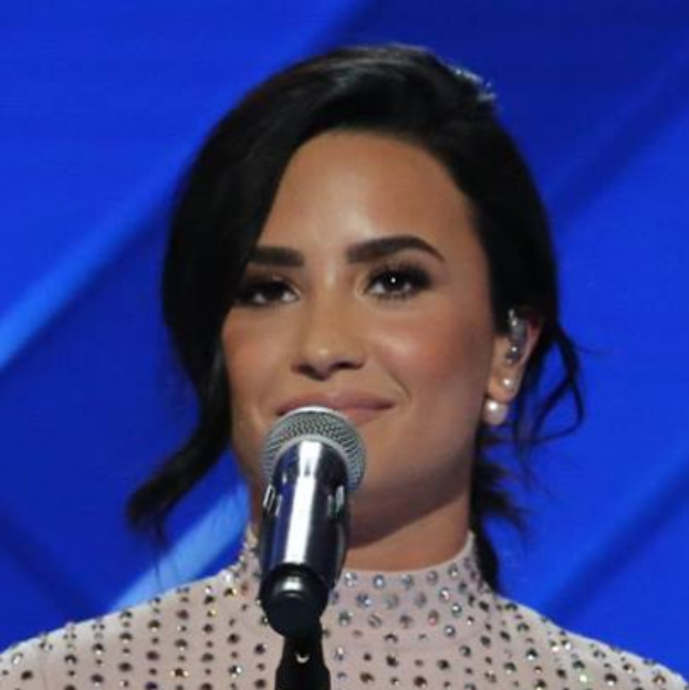 Demi Lovato, in einem schwarzen Kleid und einem stylischen Bob, steht selbstbewusst vor einem blau beleuchteten Grammy-Mikrofon.