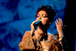 Eine Frau in einem goldenen Kleid singt in ein Mikrofon vor einem blauen und schwarzen Hintergrund, mit Eurovision Song Contest-Text unten.