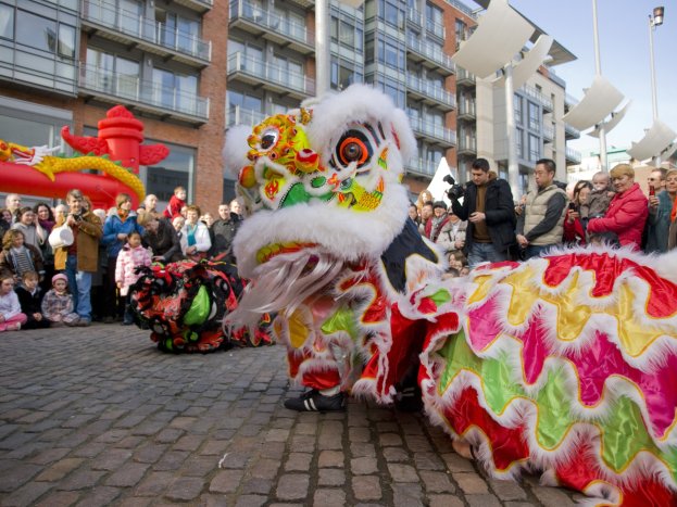 Ein farbenfrohes chinesisches Neujahrsfest in Amsterdam mit einem Löwen tanzen vor einer Zuschauermenge, einige halten Kameras, vor einem Hintergrund aus Gebäuden, Laternenmasten und einem klaren blauen Himmel.