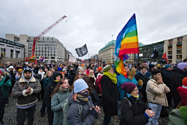 Große Gruppe von Menschen bei der LGBTQ+-Rechte-Demo in Berlin, mit Fahnen und Plakaten, vor einem Gebäude mit einem Kran und einem bewölkten Himmel im Hintergrund.