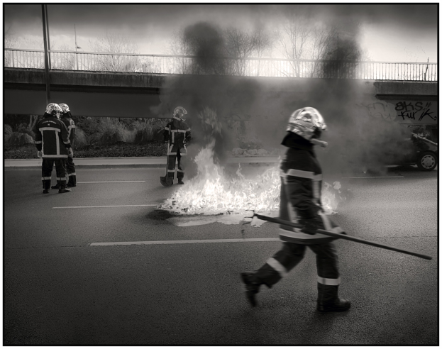 Schwarzes und weißes Bild von Feuerwehrleuten in Helmen mit Werkzeugen, die vorbei an einem Feuer gehen, mit einem Fahrzeug, Brücke, Bäumen, Pfählen und einer Wand mit Graffiti im Hintergrund.