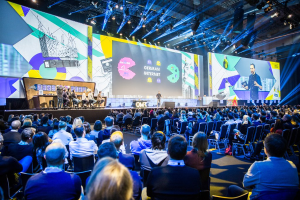 Eine große Menschenmenge, die auf Stühlen vor einer Bühne sitzt, mit einigen Personen, die auf dem Podium im Vordergrund stehen, einem Tisch mit verschiedenen Gegenständen links und Bildschirmen und Lichtern im Hintergrund auf der Deutschen Internet-Konferenz 2019.