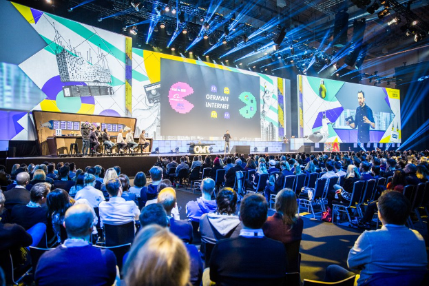 Eine große Menschenmenge, die auf Stühlen vor einer Bühne sitzt, mit einigen Personen, die auf dem Podium im Vordergrund stehen, einem Tisch mit verschiedenen Gegenständen links und Bildschirmen und Lichtern im Hintergrund auf der Deutschen Internet-Konferenz 2019.