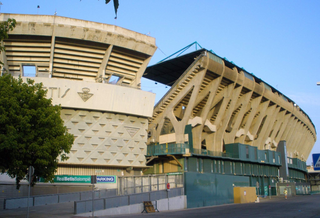 Großes Stadion mit zahlreichen parkenden Autos davor, umgeben von Gebäuden, Bäumen, Strommasten, Schildern, Geländern und einer Straße, unter einem sichtbaren Himmel.