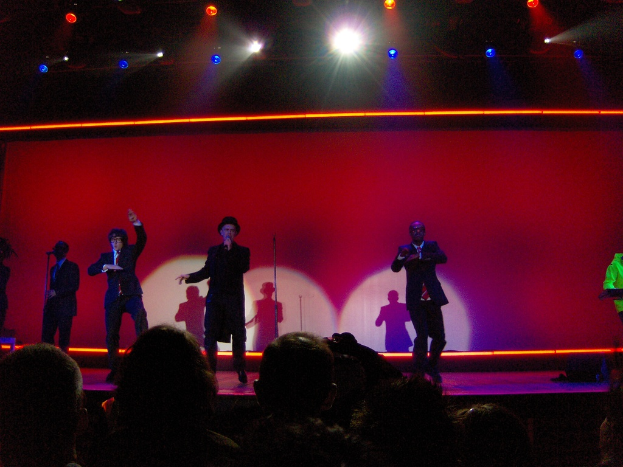 Ein Mann in einem schwarzen Kleid hält ein Mikrofon auf der Bühne, neben ihm tanzen drei Tänzer; ein Kameramann und Zuschauer sind unten, und ein Mann in einer grünen Jacke spielt rechts am Piano, während Scheinwerfer von oben leuchten.
