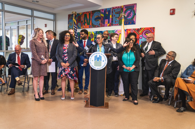 Gruppe von Menschen vor einem Rednerpult mit Mikrofon und einer Tafel mit Text, der "Sancho-Stadtrat tritt dem Stadtrat bei" anzeigt, mit sitzenden und stehenden Teilnehmern, Flaggen, Feuerlöscher und Deckenbeleuchtung im Hintergrund.